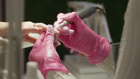 close-up of nail technician in pink gloves shaping client nail using electric file while wearing face mask. client wears gold ring. technician s face partially visible with soft background blur