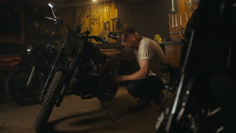 confident blond guy in a white t-shirt mechanic works and repairs a motorcycle in his workshop