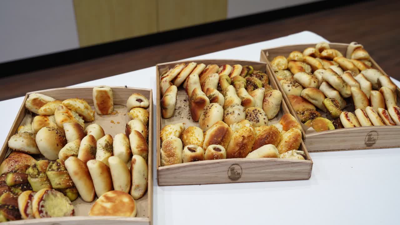 Wide shot of assorted breadsticks and mini pastries arranged in wooden trays on a buffet table indoors, showcasing bakery catering selection under bright lighting