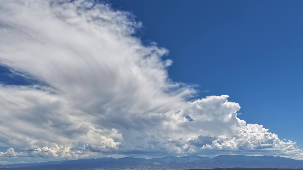 4K drone timelapse of a massive, elongated cloud formation stretching across a vibrant blue sky. The huge white clouds loom over the distant Sangre de Cristo mountain range on a clear summer day.