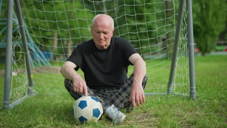 an elderly man sits on the grass near a goal post, gently touching a soccer ball with his right hand, with trees in the background