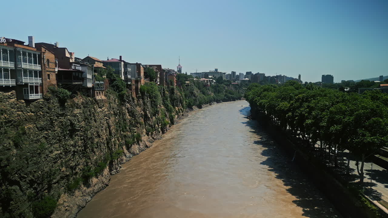 Panoramic view of the Mtkvari River flowing through Tbilisi, Georgia, with historical buildings on cliffs and green trees along the banks