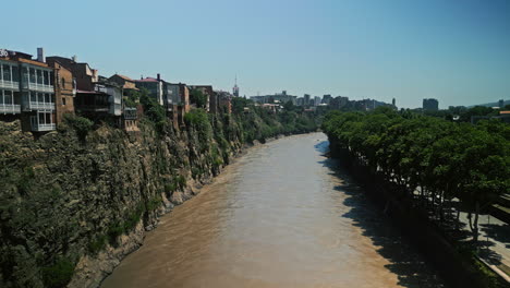 panoramic view of the mtkvari river flowing through tbilisi, georgia, with historical buildings on cliffs and green trees along the banks