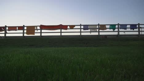 farm land in a scene, dark green grass, wooden farm fence with clothes drying on it. rags in white and orange colors. blades of grass in a foreground. evening time, sun has set. filmed in hd
