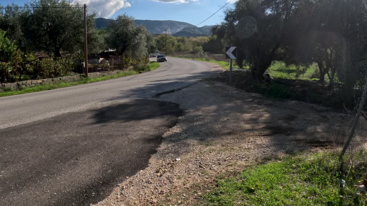 Carros andando em estrada rural de asfalto sinuosa na zona rural de Corfu, cercada por oliveiras e montanhas em um dia ensolarado