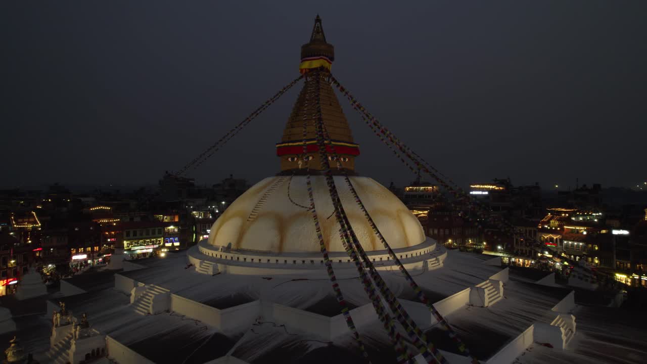 A calm night drone view of Boudhanath Stupa, with boudha and nath noted separately, glowing above Kathmandu. The sacred Buddhist landmark stands radiant in the quiet evening cityscape