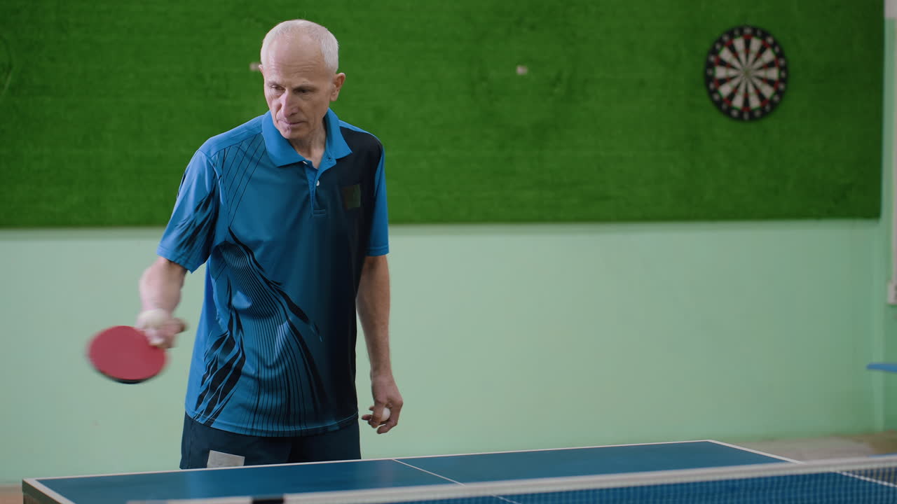 Elderly man in blue sports shirt plays table tennis indoors, preparing for serve with paddle in hand, showing discipline, and determination while training with others at indoor practice session