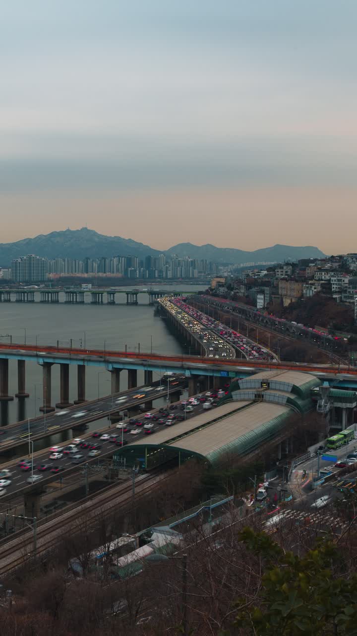 Un timelapse verticale cattura il denso traffico dell'ora di punta al tramonto che si snoda lungo l'autostrada Gangbyeonbuk-ro vicino alla stazione di Oksu, con i ponti del fiume Han e lo skyline montuoso di Seul
