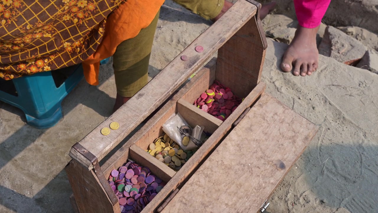 High-angle shot of a wooden box with color-coded tokens representing different brick haul values at a Bangladesh yard. Documents variable piece-rate wage systems and traditional labor tracking.