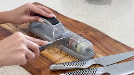 hands use a manual knife sharpener on a wooden cutting board in a bright kitchen, demonstrating proper knife sharpening technique with multiple knives visible