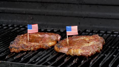 two juicy rib-eye steaks sitting on the grill and cooking a hand comes in and sticks a tiny american flag with a tooth pick into one of them