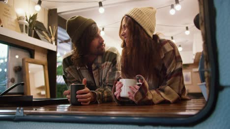 Close-up-shot-of-a-happy-brunette-guy-in-a-Green-hat-in-a-Green-checkered-shirt-communicates-with-his-brunette-girlfriend-in-a-pink-checkered-shirt-and-they-hold-mugs-with-a-hot-drink-during-their-vacation-in-a-trailer-in-a-camp-during-a-picnic-outside-the-city