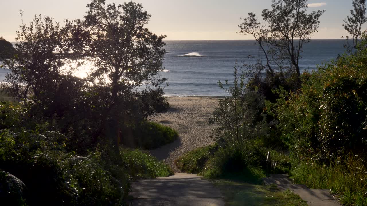Slow motion landscape of ocean waves breaking on sandy beach bay shoreline with bushland trees and concrete walking pathway driveway at Bateau Bay Central Coast suburbs tourism travel nature Australia
