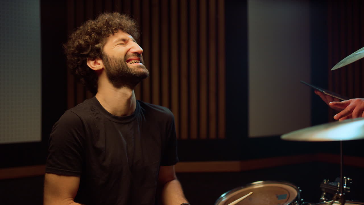 un músico alegre riendo en el estudio, un baterista sonriente hablando en el pasillo.