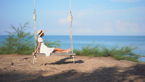 a young woman in a white sundress rocks back and forth in a suspended swing while looking out on the ocean horizon