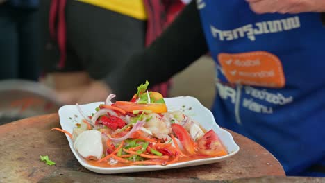 plating a spicy seafood somtam salad, a mixture of cuttlefish, squid, tomatoes, onions, celery, carrots, lime, fish sauce, coconut sugar, with a dash of salt and pepper flakes