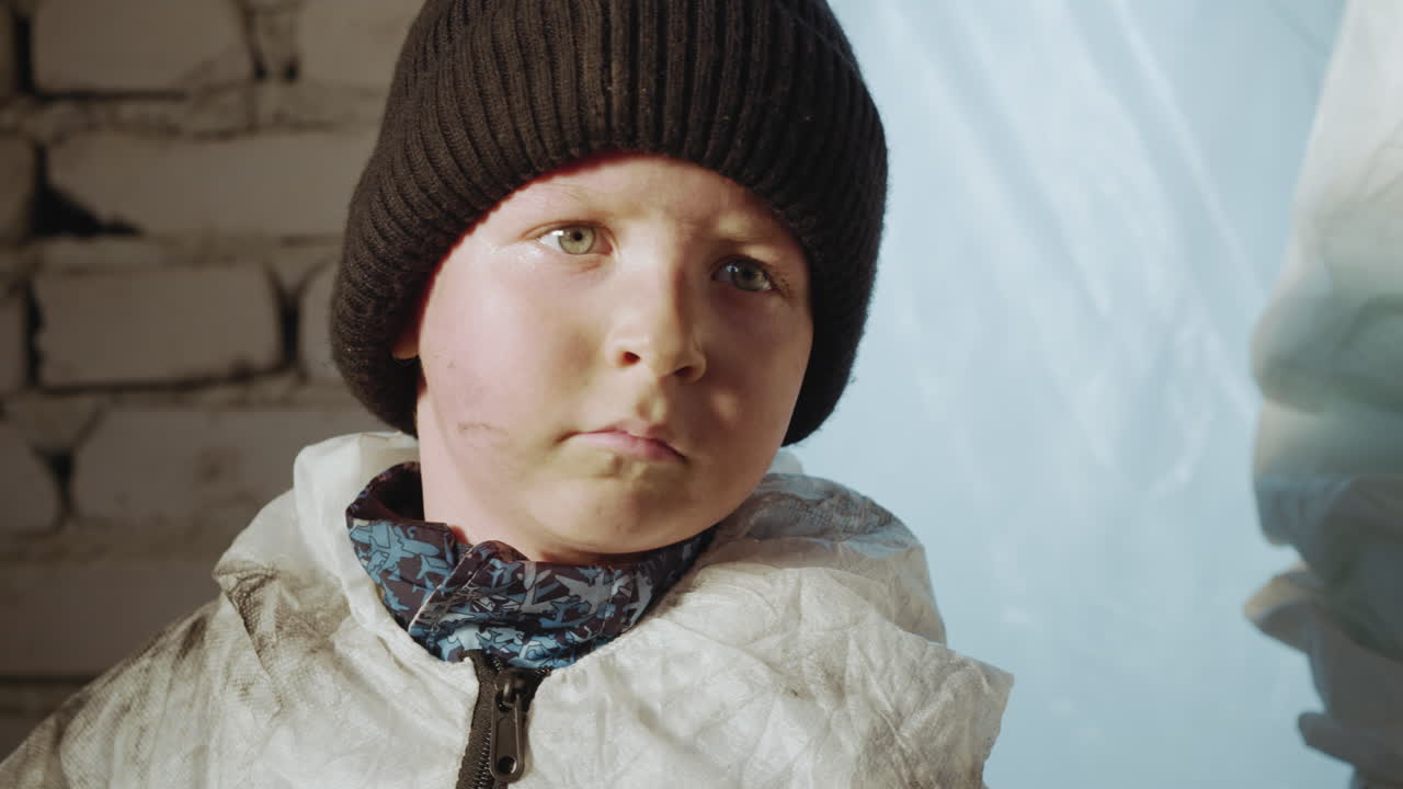 Close up of serious young boy wearing dark beanie and white protective suit with smudges, standing indoors near painted brick wall, turning slightly with focused expression in soft natural light