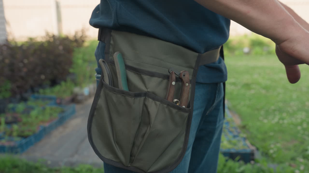 Close side view of gardener with waist apron retrieving tools, sliding into pocket while utility knife slips from hand, outdoor nursery background with green plants
