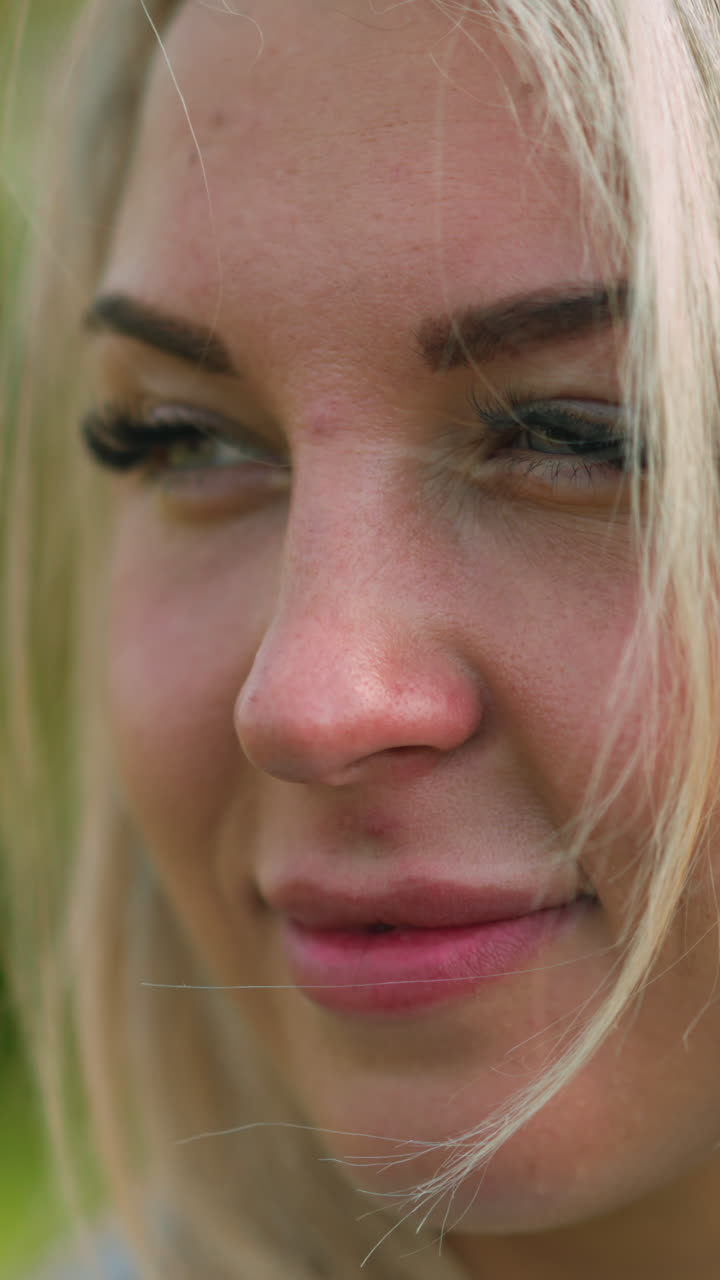 Attractive woman with long blonde hair and fake eyelashes smiles for camera standing on blurred green background at light wind closeup slow motion