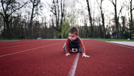 caucasian baby boy sits on the track at the stadium with a football ball. kid stands up shaking his hands.