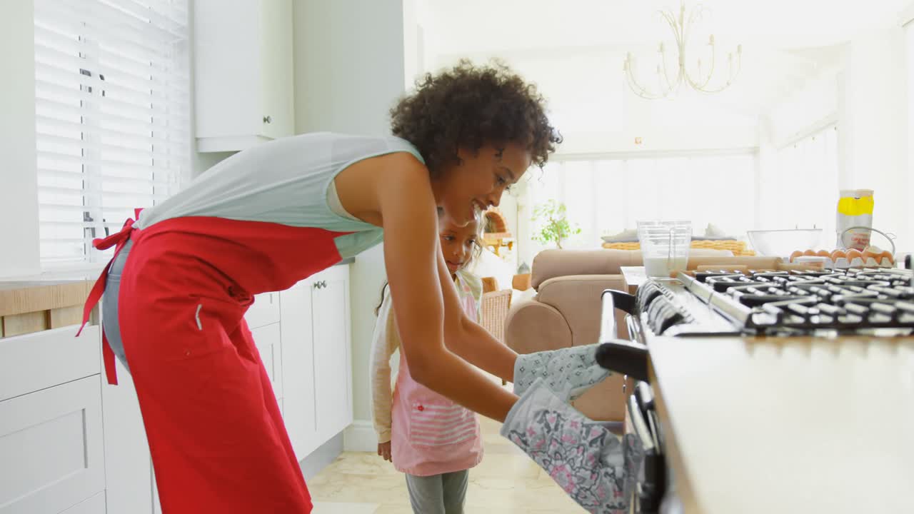 Side view of black mother and daughter using oven machine in kitchen at home 4k