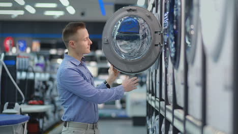 A-young-handsome-man-chooses-a-smart-washing-machine-in-a-consumer-electronics-store-for-his-new-home.-Studies-the-characteristics.