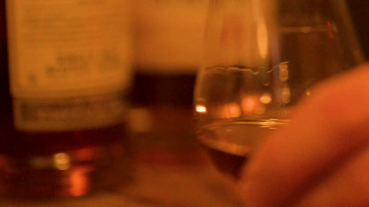 A hand lifts whisky tasting glass from bar, amber liquid, warm evening lighting, shallow focus