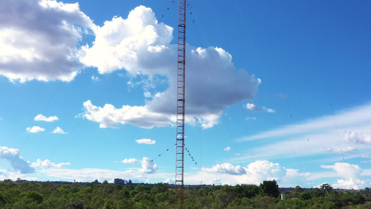 Mastro de antena vertical com escoras diagonais e faixas de aviso ergue-se contra o cenário de nuvens cúmulos, com densa floresta de cerrado verde cercando a elevação da base