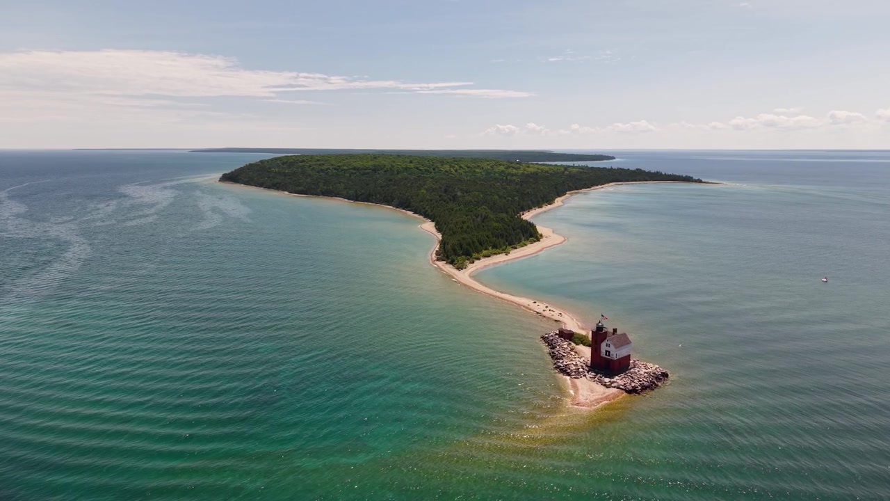 Široké dronové panorama zalesněného ostrova Round Island s písečnými břehy a majákem zasahujícím do klidných vod Velkých jezer poblíž Hessel v Michiganu.
