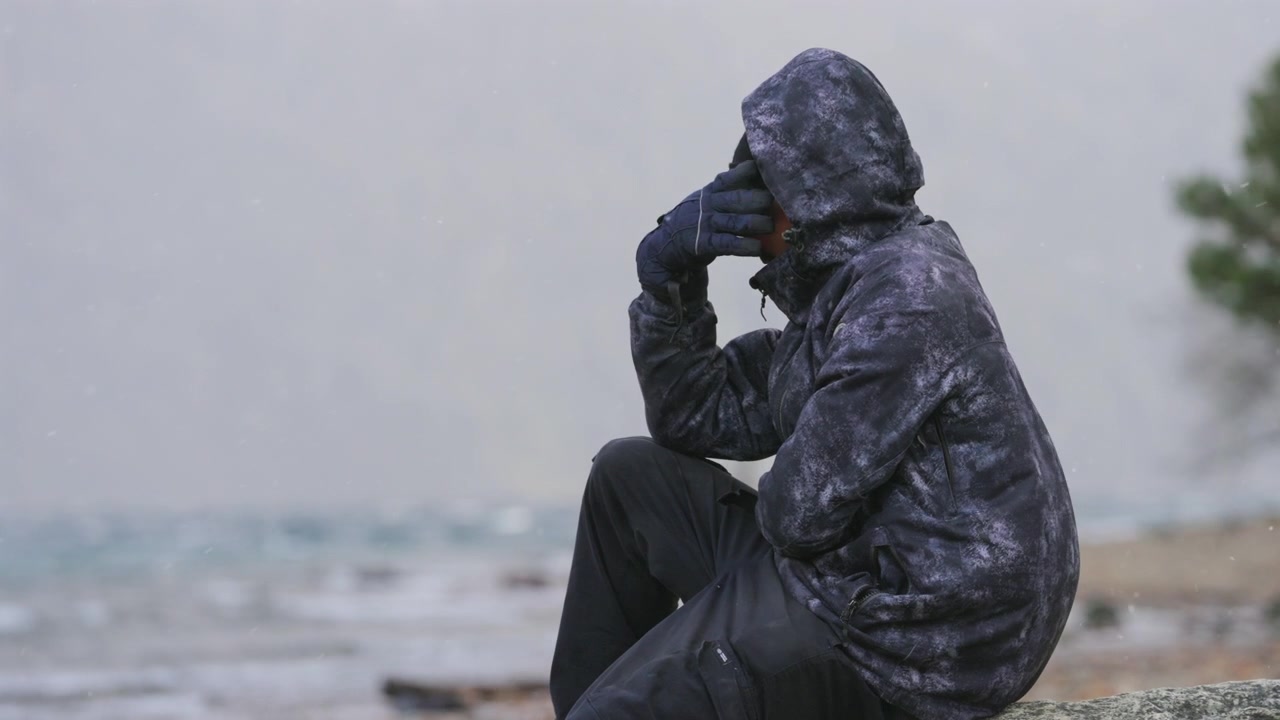 A man in a hooded jacket sits by the shoreline, appearing deep in thought. The overcast sky and cold weather create a somber atmosphere. Suitable for themes of contemplation or solitude.