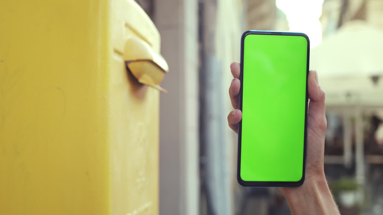 Close-up of a person holding a smartphone with a green screen while another person interacts with a yellow public box