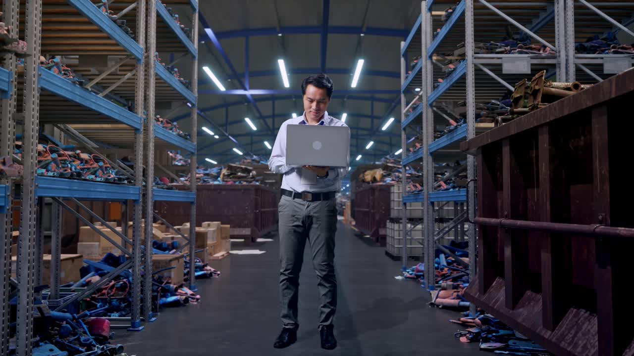 Figura intera di un lavoratore professionista asiatico in piedi con il suo laptop, all'interno di un magazzino industriale di rottami metallici e riciclaggio, mentre lavora ininterrottamente.