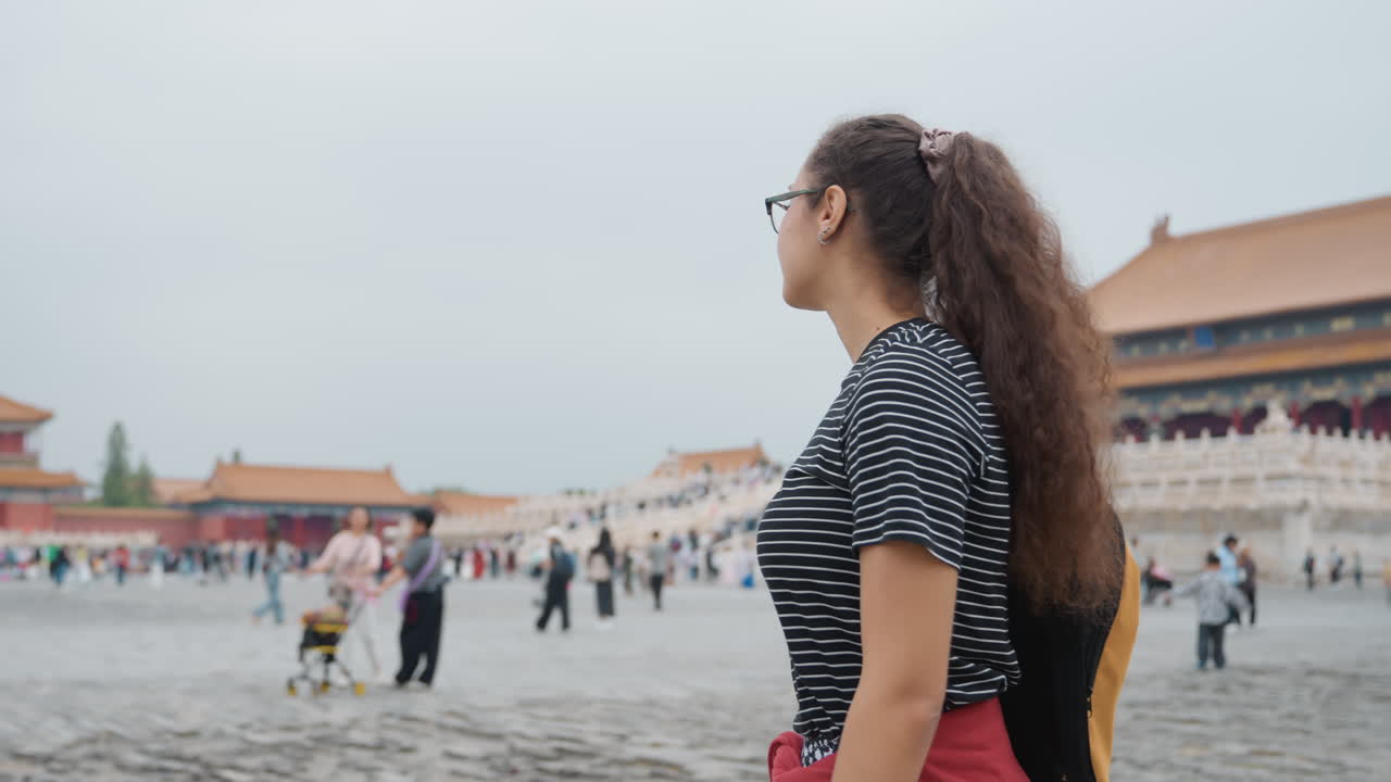 Una giovane donna di colore visita il palazzo di Pechino, una viaggiatrice solitaria si ferma in un ampio cortile, osservando lo skyline e un tempio storico, con uno zaino in spalla, cielo coperto, folla che si aggira, umore contemplativo