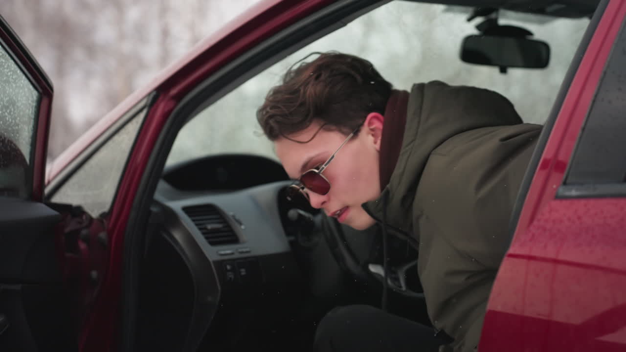 Man wearing winter jacket exits red car during cold weather with visible snowflakes in air and water droplets on vehicle, capturing moment of winter activity in outdoor snowy setting