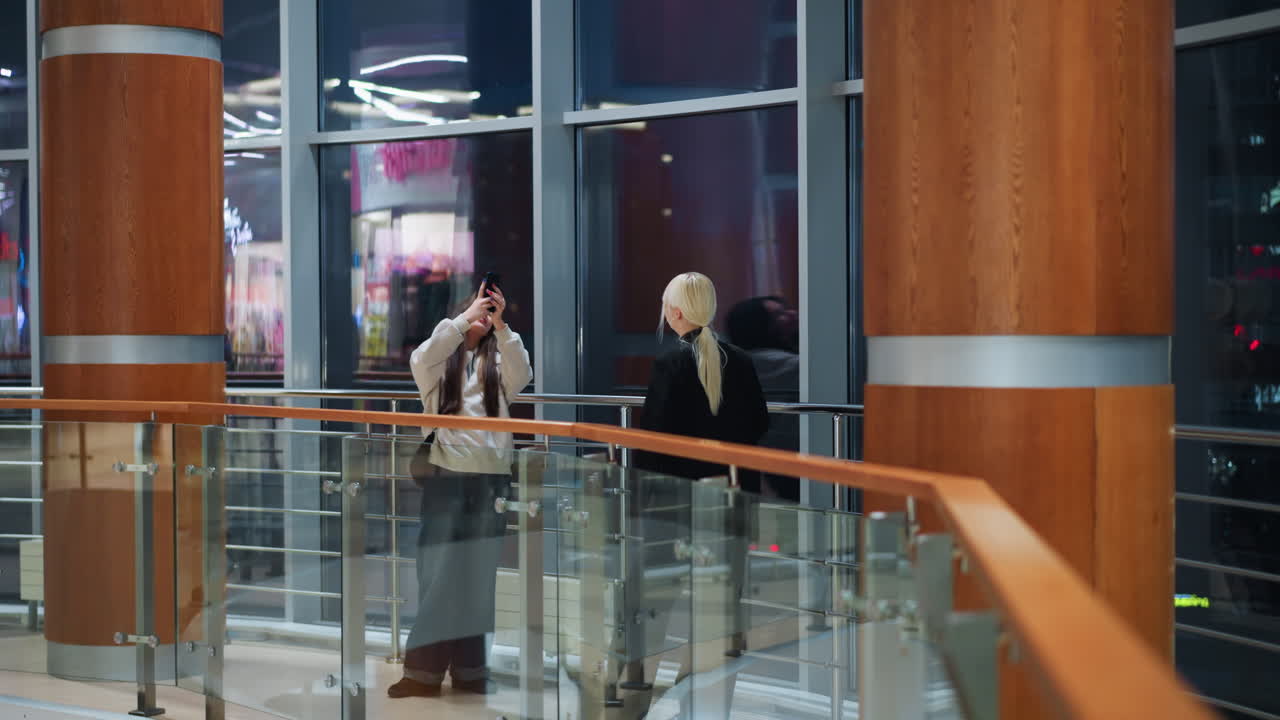 Girl with long hair holding phone taking photo of her sister indoors near large glass window while colorful city lights blink and reflect on glass inside modern shopping mall