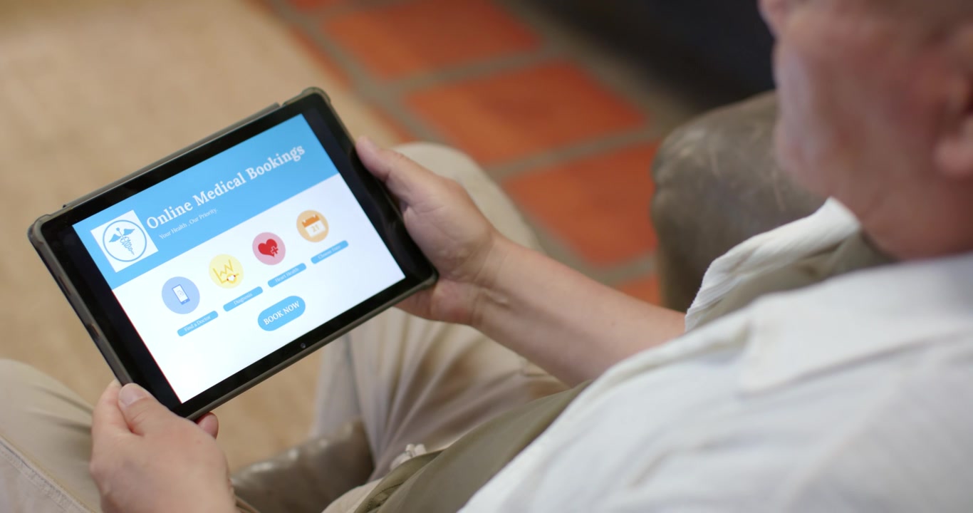 Senior man holding tablet prompted by booking button, adjusting grip while booking visit on couch