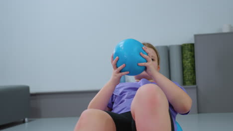 niña joven equilibrándose en una pelota de estabilidad azul mientras se acuesta de espaldas, levantando las manos en un movimiento controlado, involucra los músculos del núcleo mientras gira en la pelota de ejercicio
