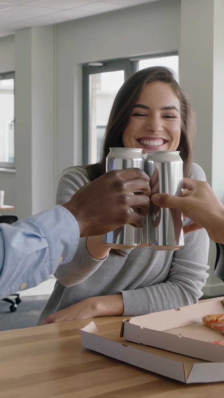 Video vertical: Brindis con latas después de que el equipo las levanta; ella, de gris, celebra en la oficina con pizza.