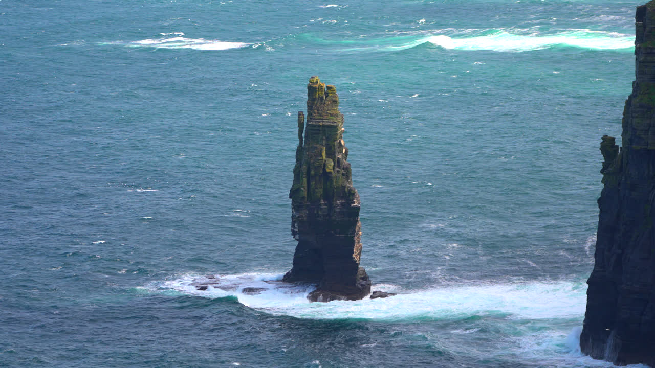 A massive sea stack stands tall in the waters near the Cliffs of Moher, with steep cliffs in the background. Waves crash against the rock, highlighting the dramatic and wild Irish coastline.