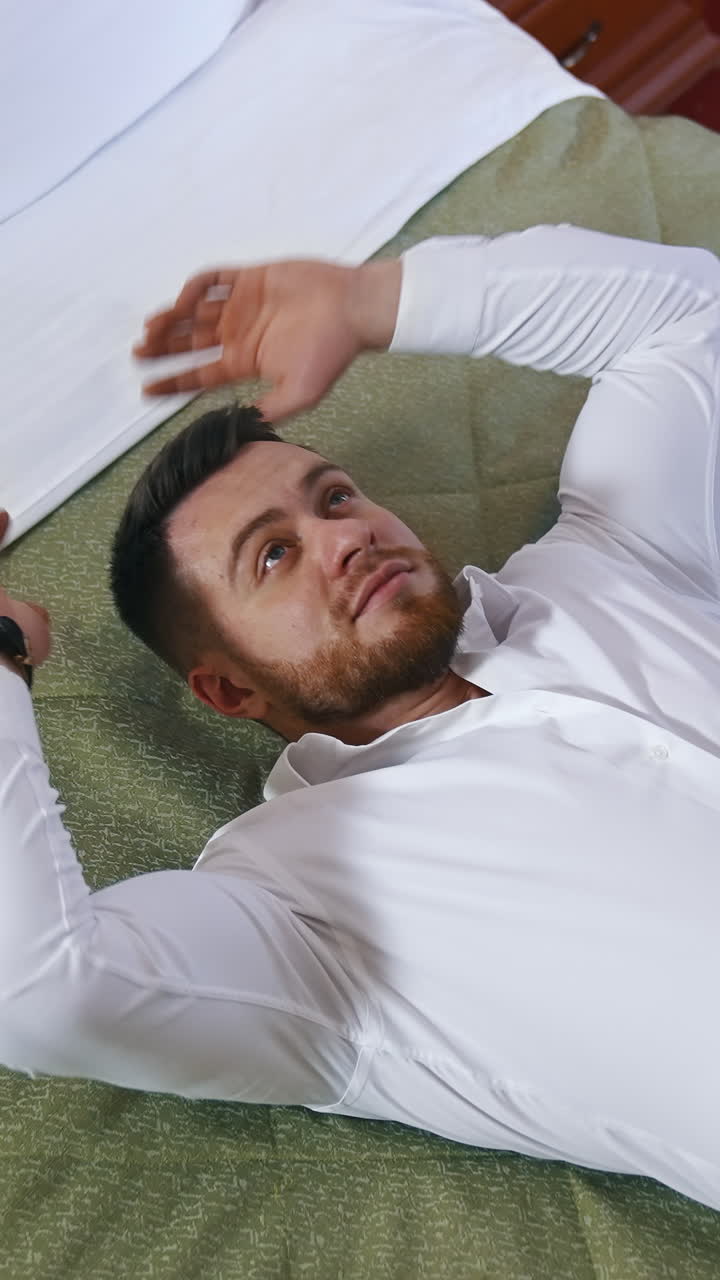 Young manager lays on a bed in a hotel room. View from above on a tired businessman after long trip. Modern interior of a hotel room. Vertical video