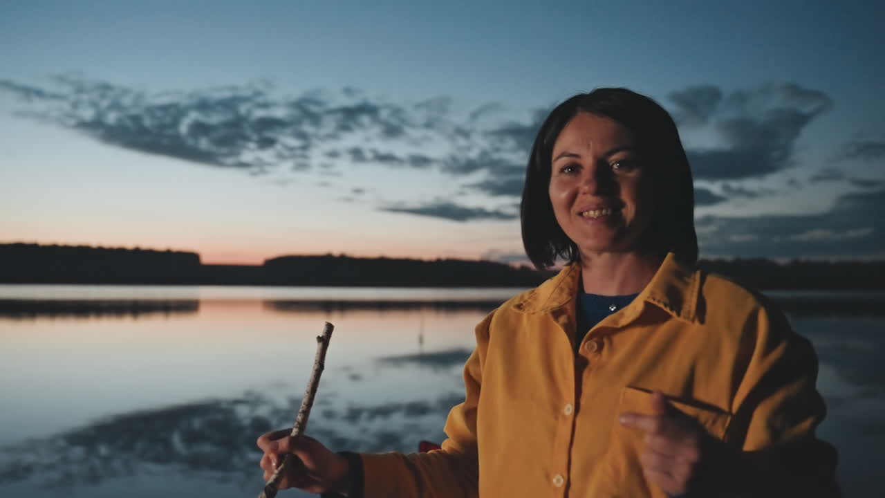 Camperista serale in riva al lago sorride arrostendo qualcosa con un bastone. Una donna caucasica ride mentre tiene un bastone per arrostire, momento implicito di marshmallow. Caldo bagliore e luce delle braci, energia narrativa, atmosfera accogliente da campeggio.