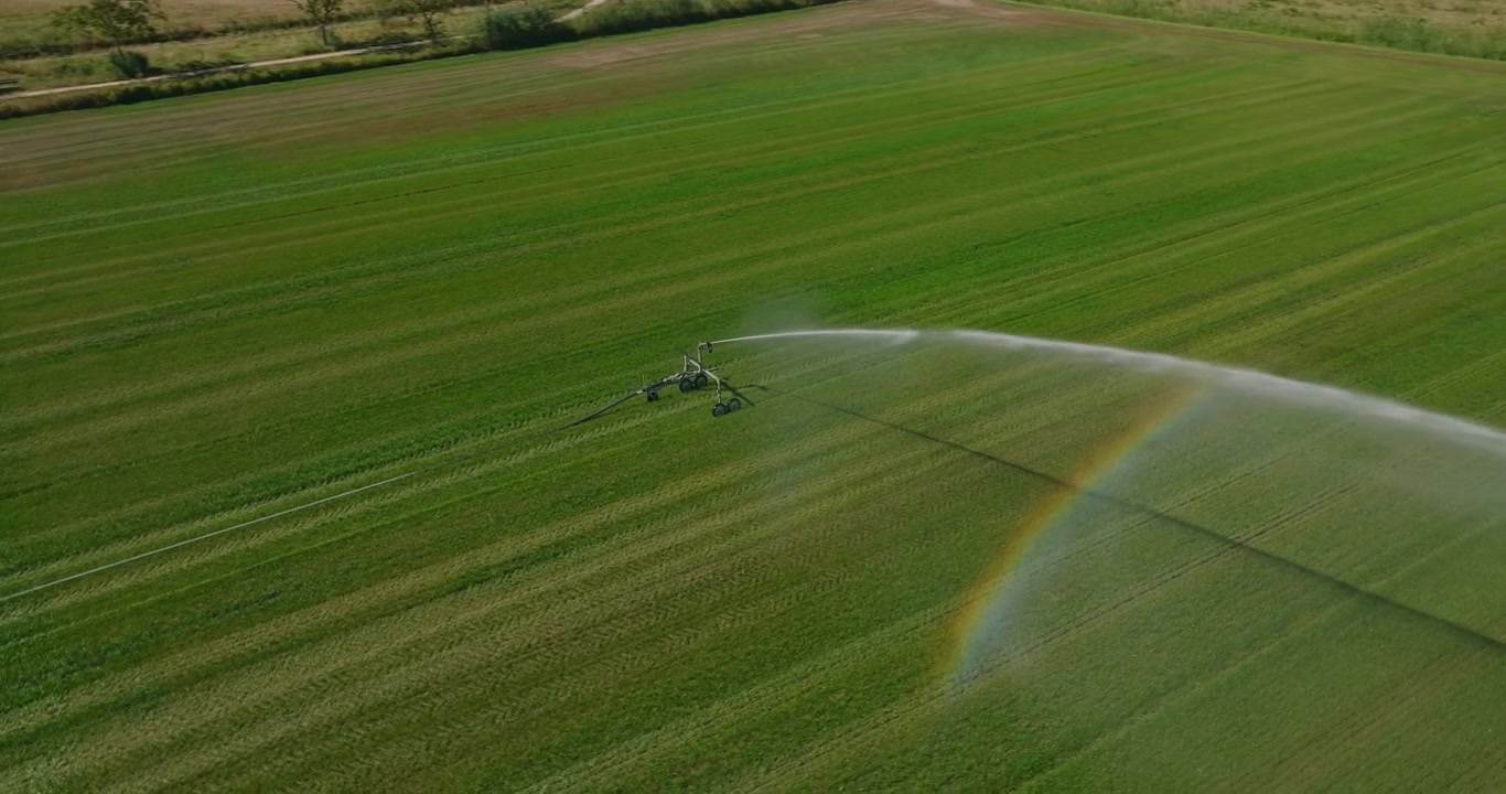 Un drone orbita un potente irrigatore che annaffia un campo agricolo verde in una giornata di sole, creando un arcobaleno magico attraverso l'arco bianco dell'acqua nebulizzata.