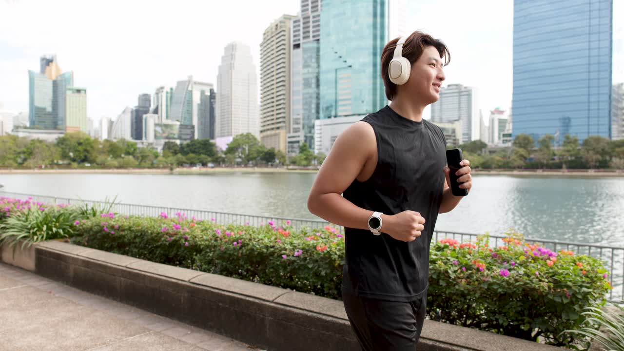 Seorang pria Asia yang bugar berlari di sepanjang jalan setapak taman sambil mengenakan headphone dengan latar cakrawala kota