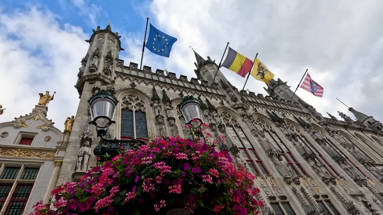 Low-angle view of European, Belgian, and city flags waving atop ornate medieval city hall with vibrant flowers, under bright natural daylight and partly cloudy sky