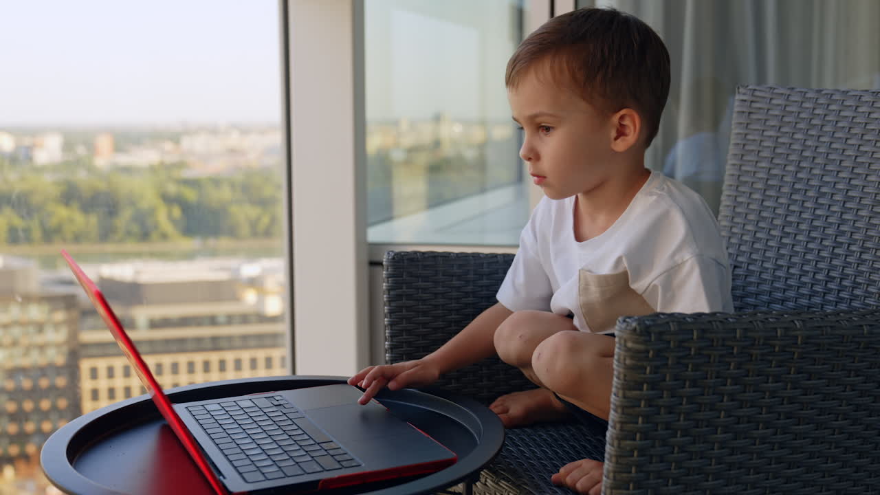 Dark-haired Caucasian toddler boy sitting in the armchair using laptop. Little kid uses a touch pad