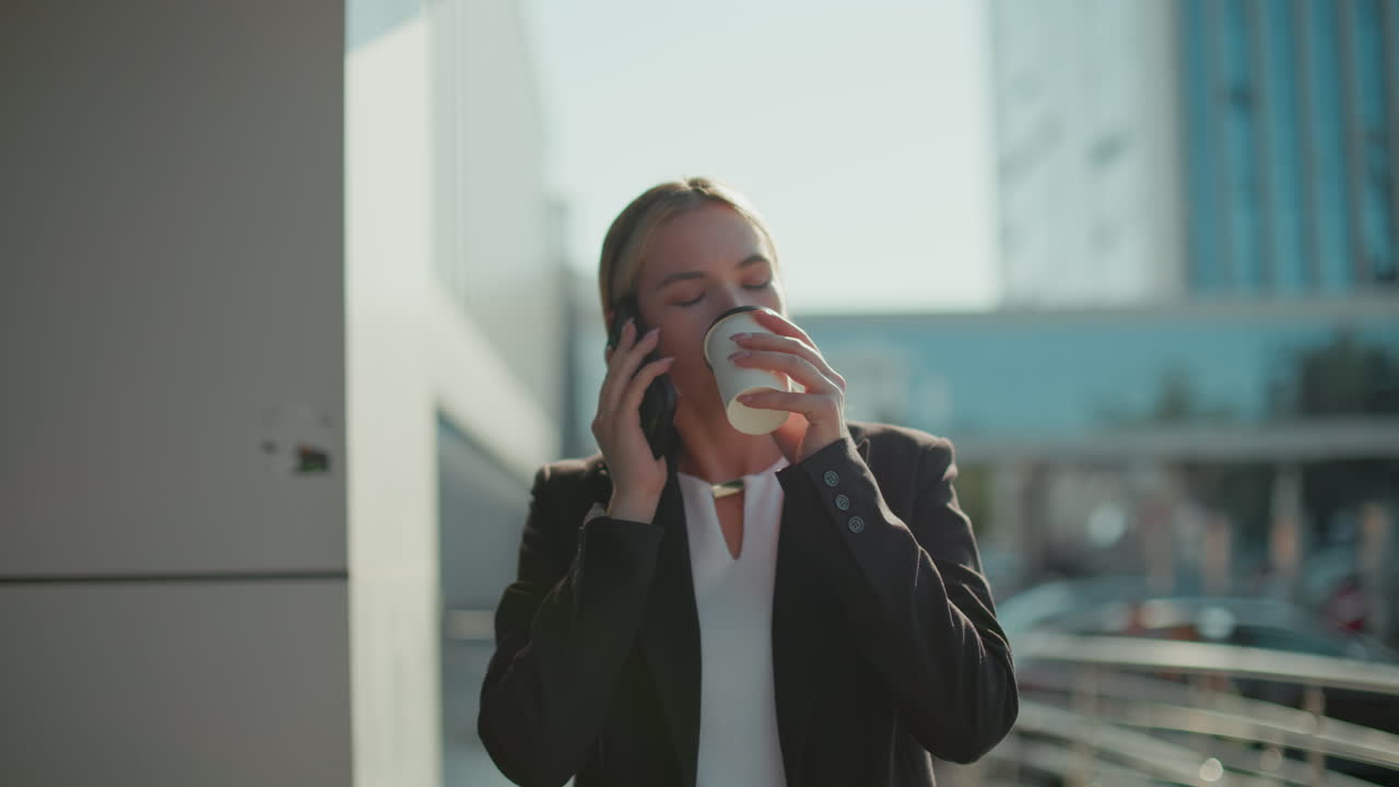 White lady on call drinking coffee as she walks by office building in professional attire, with blurred parked cars in background, sunlight reflecting her face