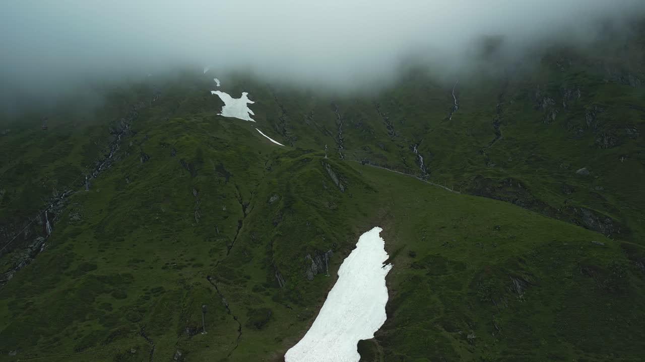 montañas verdes con parches de nieve y cabinas de teleféricos rojos entrando y saliendo de las nubes