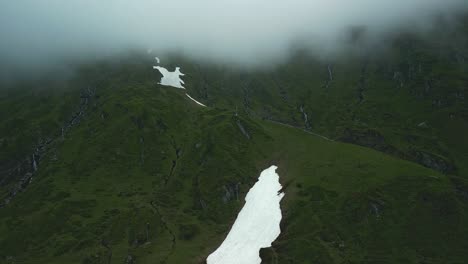 Grüne-Berge-Mit-Schneeflecken-Und-Rote-Seilbahnkabinen,-Die-In-Die-Wolken-Hinein--Und-Wieder-Herausfahren