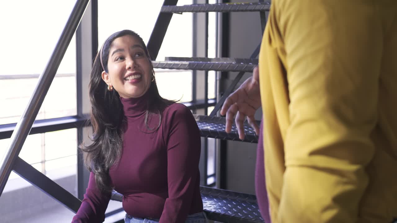 Diverse coworkers laughing on metal staircase while man sharing story beside large window