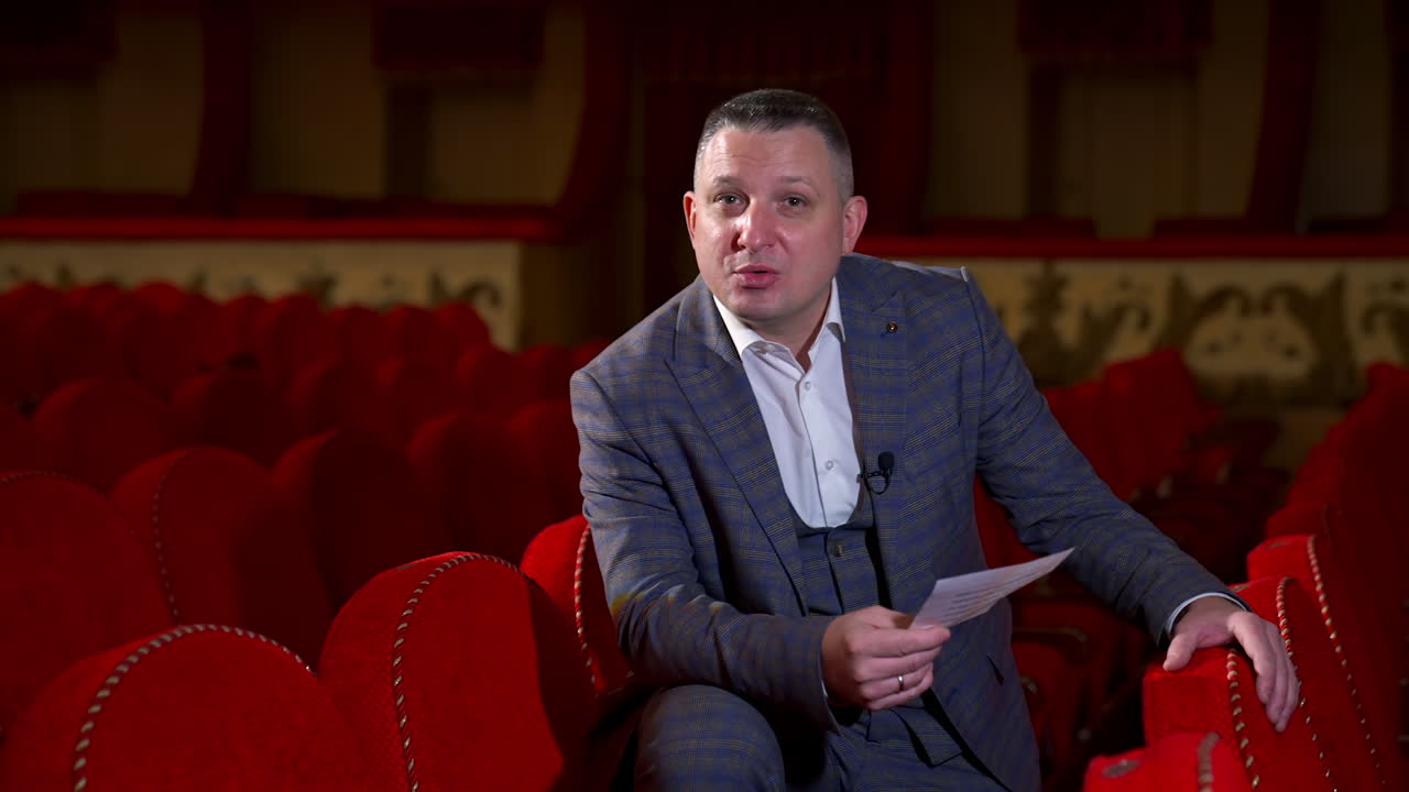 Actor at rehearsal in auditorium. Man repeating monologue while sitting among red chairs in the empty hall. Speaker rehearsing before the performance.